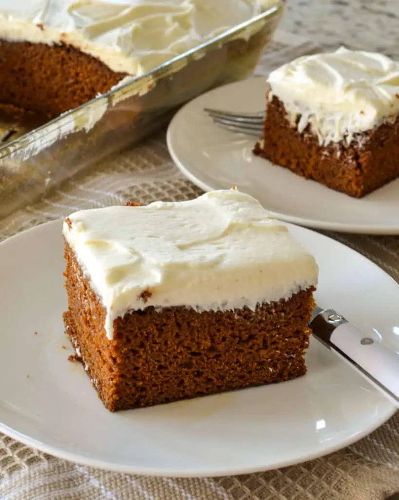 A slice of gingerbread cake with cream frosting on a single serving plate with a fork with the gingerbread cakein the background. 