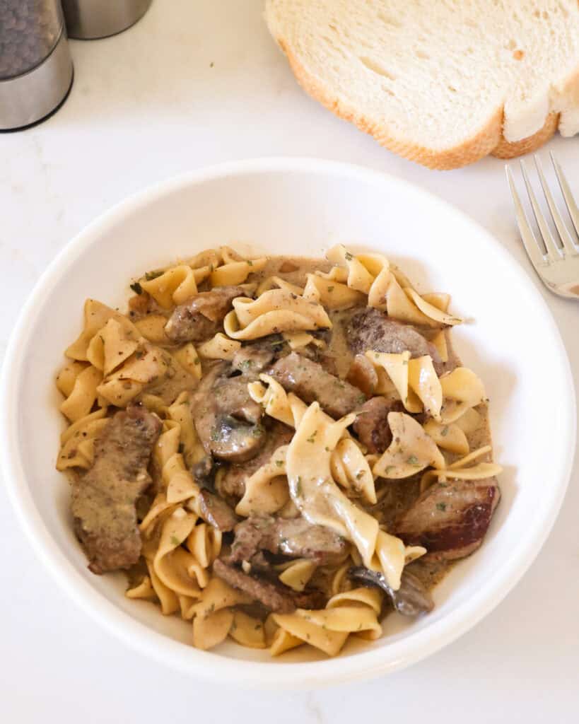 Beef stroganoff in a single serving bowl with bread and salt and pepper shaker in the background. 