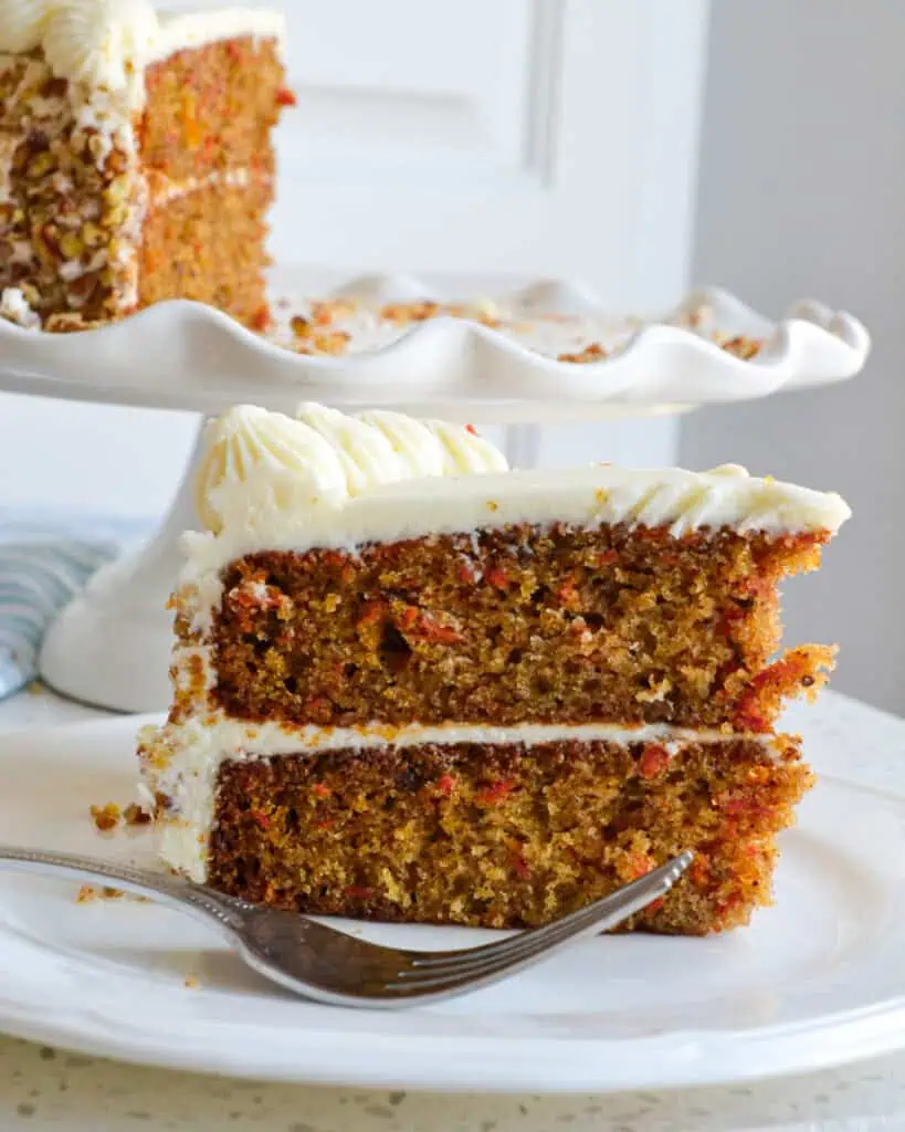 The side view of a slice of carrot cake on a plate with a fork in the foreground. 