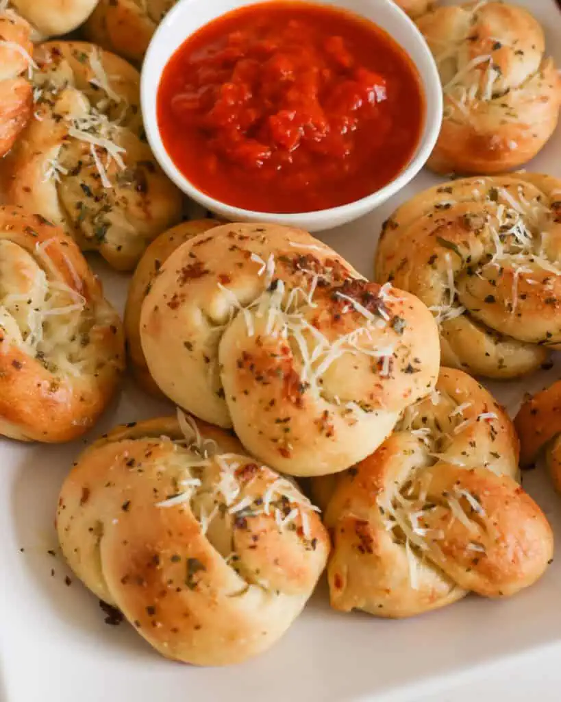 A close up view of warm garlic knots on a serving dish with a bowl of warm marinara. 