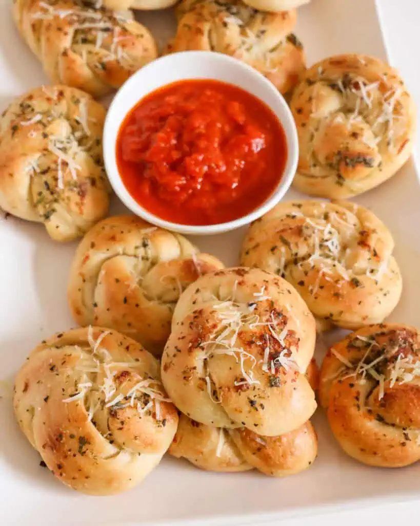 A white serving platter full of garlic knots and a bowl full of warm marinara. 