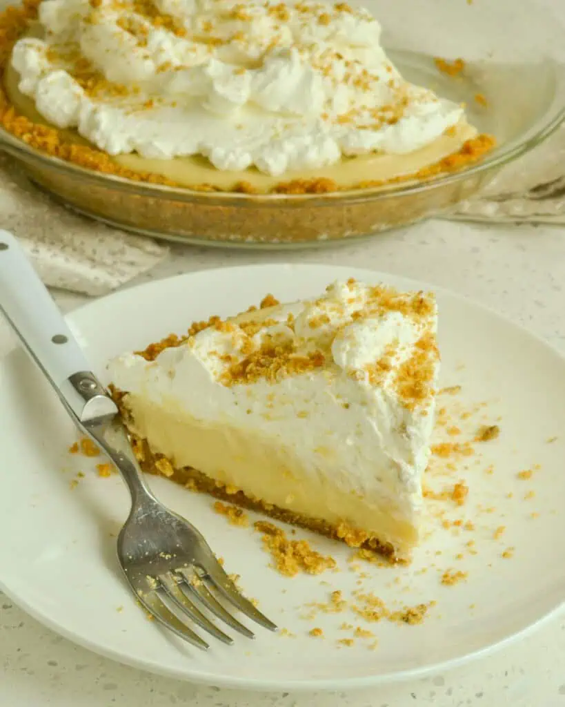 A piece of creamy butterscotch pie on a serving plate in front of the rest of the pie in the pie dish. 