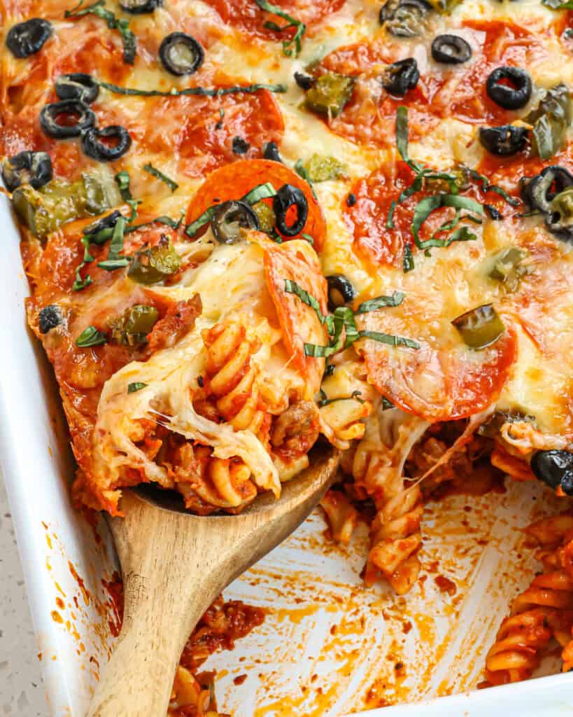 A large dish of pizza casserole with some of the casserole dished out and a big spoon in the casserole with some of the pasta on it. 