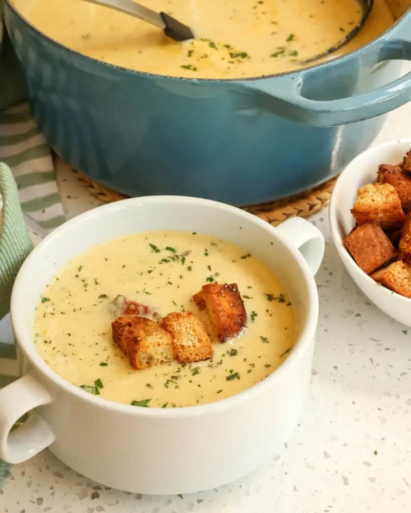 A bowl of cream of celery soup with a bowl of croutons and a Dutch oven full of cream of celery soup in the background. 