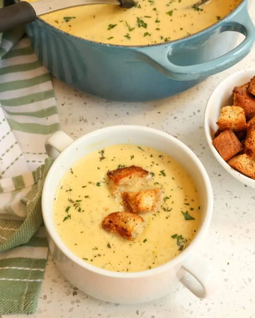 A bowl of cream of celery soup with a bowl of croutons and a Dutch oven full of cream of celery soup in the background. 