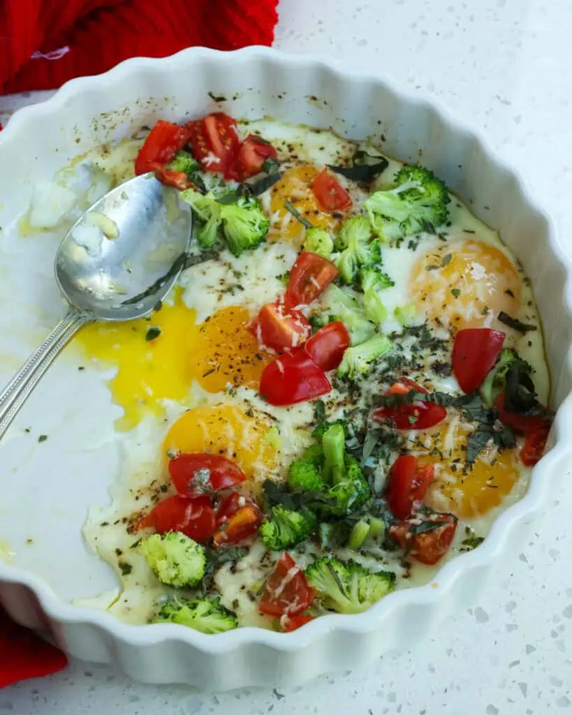 A dish full of baked eggs, tomatoes, broccoli, and Parmesan cheese with a spoon in the dish. 