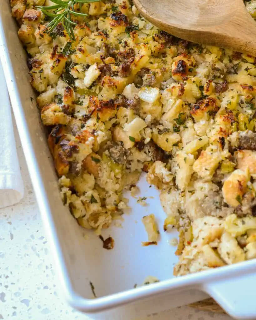 A close up view of Thanksgiving sausage stuffing with onions, celery, garlic, pork sausage, and apples, all seasoned with fresh herbs like rosemary, thyme, and sage.