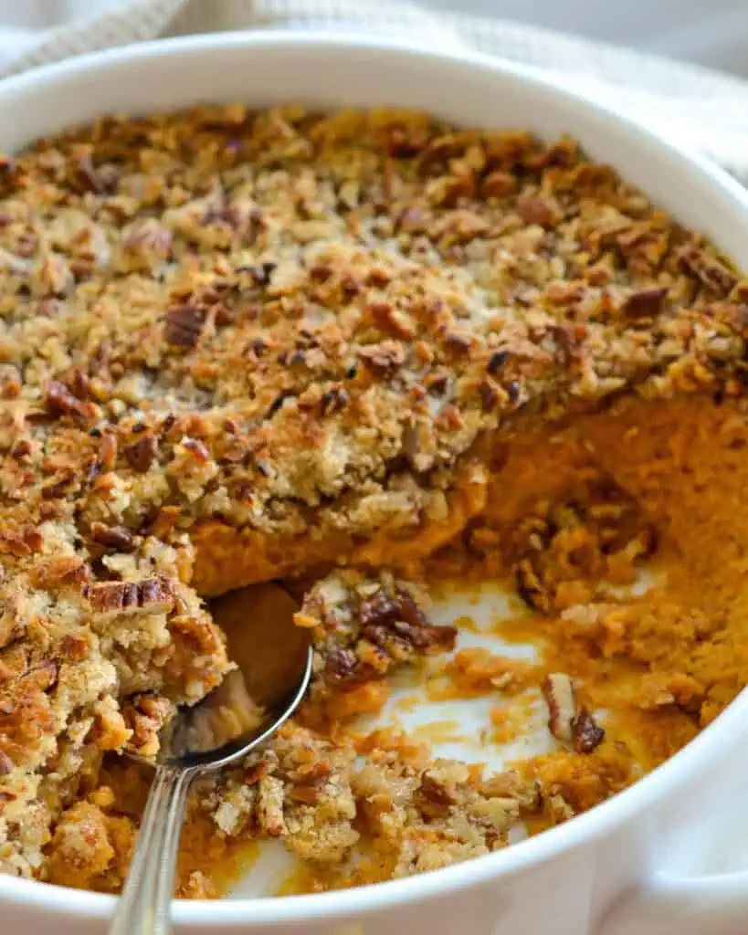 Sweet Potato Casserole with a crunchy pecan toppiing in a large round casserole dish with a spoon in it. 