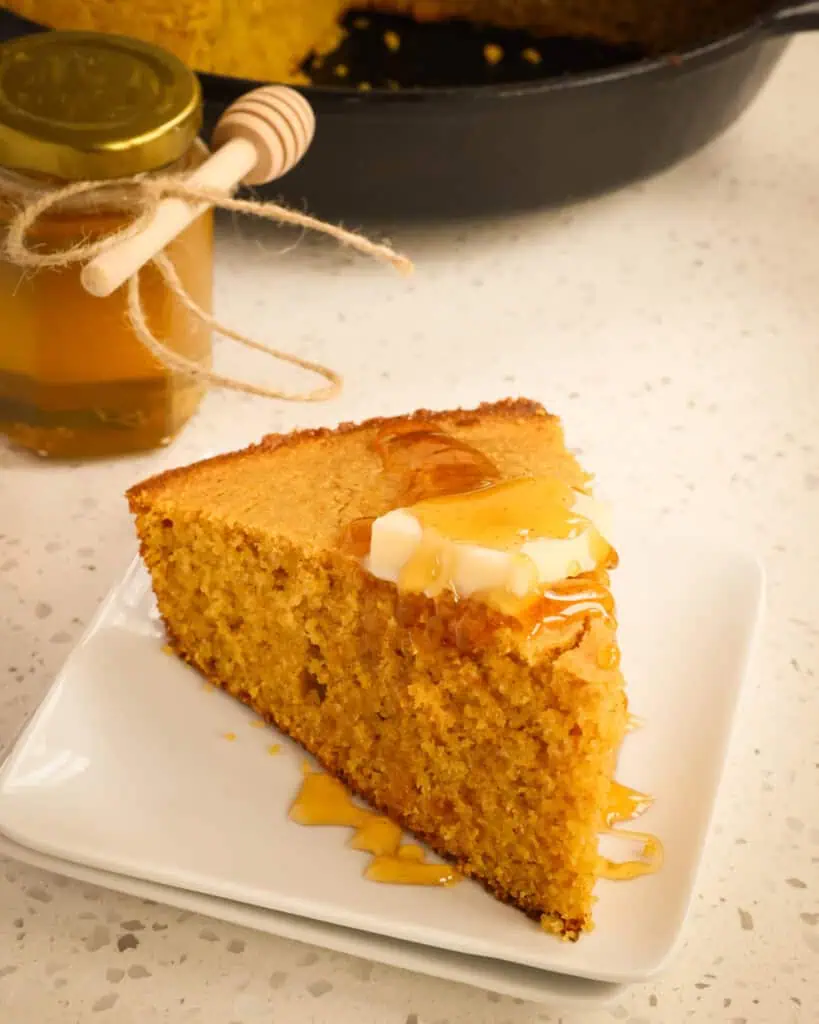 A pierce of sweet potato bread on a plate with a jar of honey and the rest of the sweet potato bread in the cast iron skillet. 