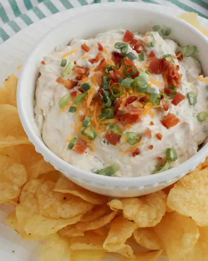 A closeup view of a bowl of crack dip garnishedwith chopped bacon, sliced green onions, and shredded cheddar cheese. 
