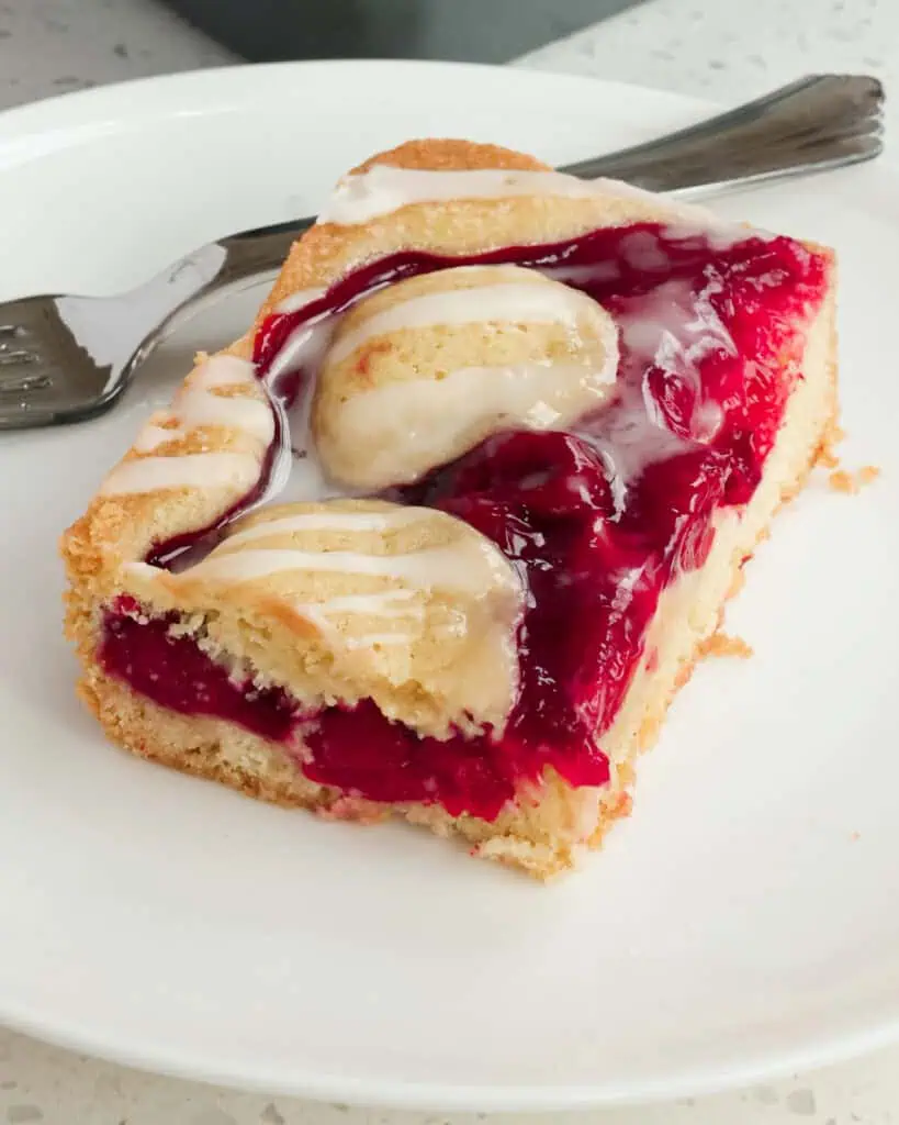 A cherry pie bar on a single serving plate with the pan of cherry pie bars in the background. 