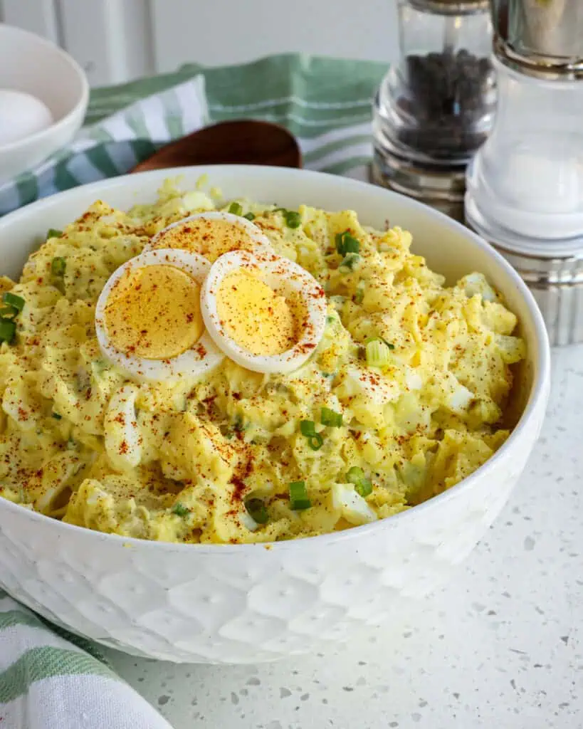 A serving bowl full of southern potato salad with salt and pepper, hard cooked eggs, and a wooden spoon the background. 