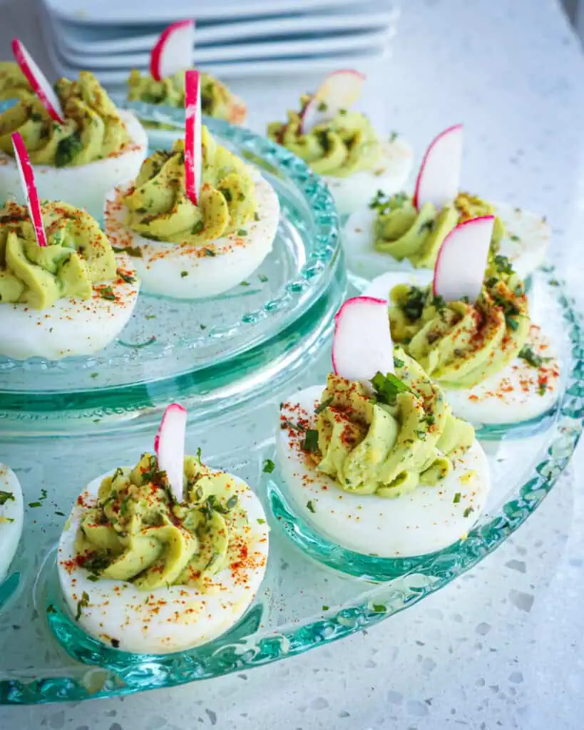 A glass egg platter full of avocado deviled eggs topped with chopped cilantro and radish slivers. 