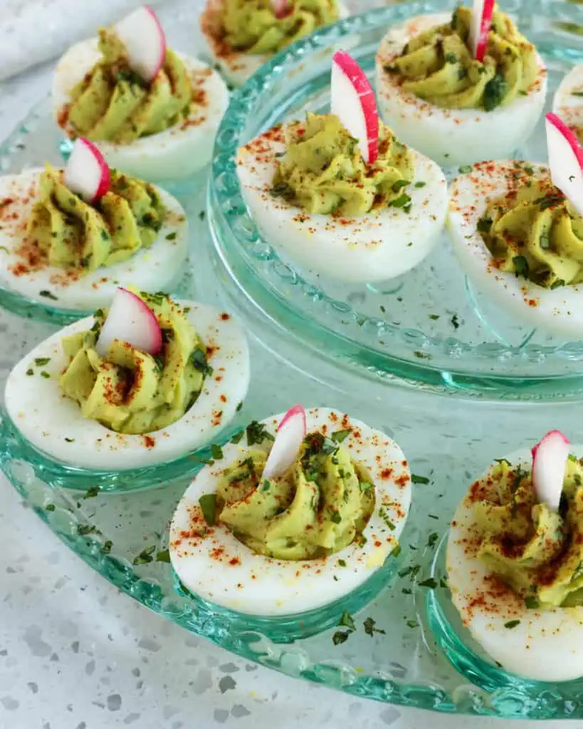 Avocado deviled eggs on a glass egg plate. The eggs are garnished with chopped cilantro and slivered radished. 