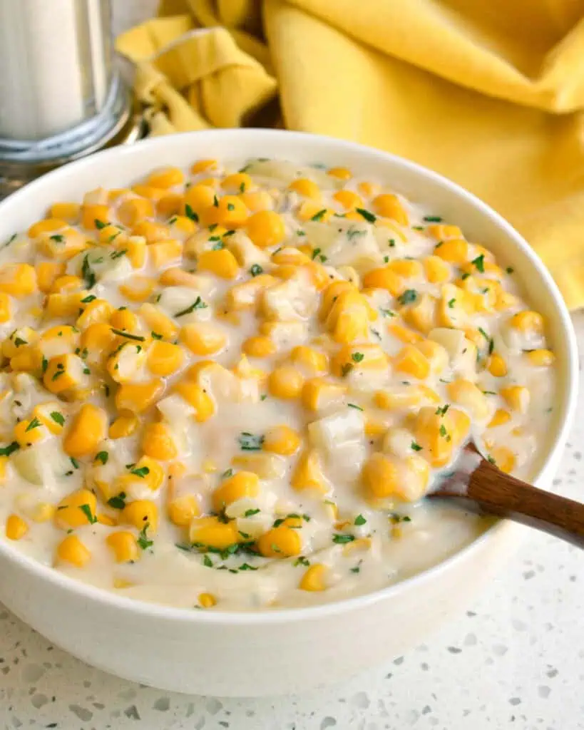 A sesrving bowl full of creamed corn with a wooden spoon and salt and pepper shaker in the background. 