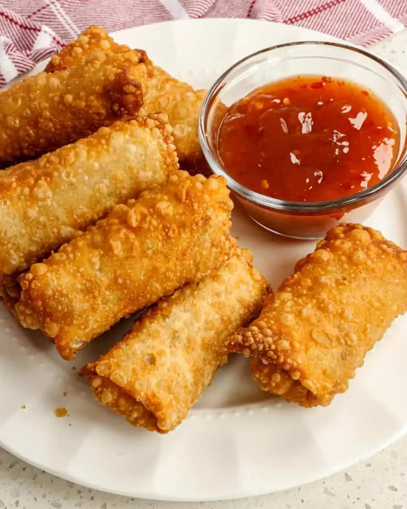 A serving plate full of homemade egg rolls with a small glass bowl full of sweet chili sauce. 