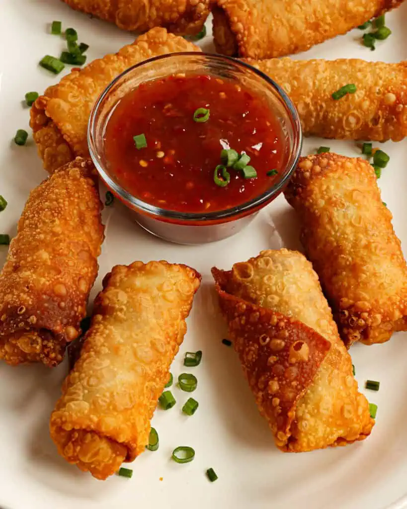 Homemade egg rolls on a large oval white platter with thinly chopped green onions with a small glass bowl full of homemade chili sauce. 