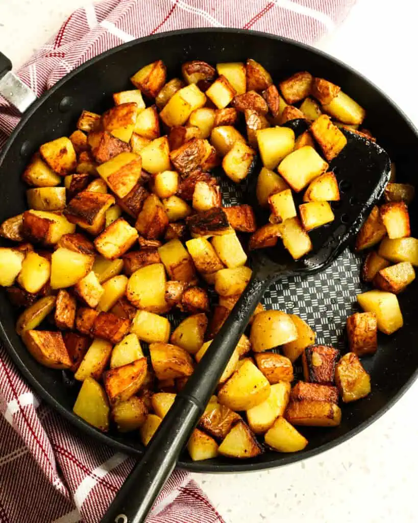 An overhead view of pan fried potatoes in a large black skillet withe a large black spatula. 