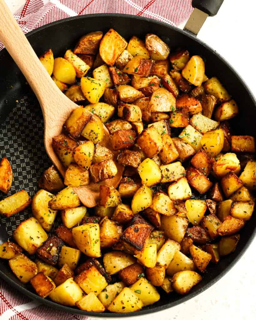 Pan fried potatoes in a large black skillet with a wooden spatula. 