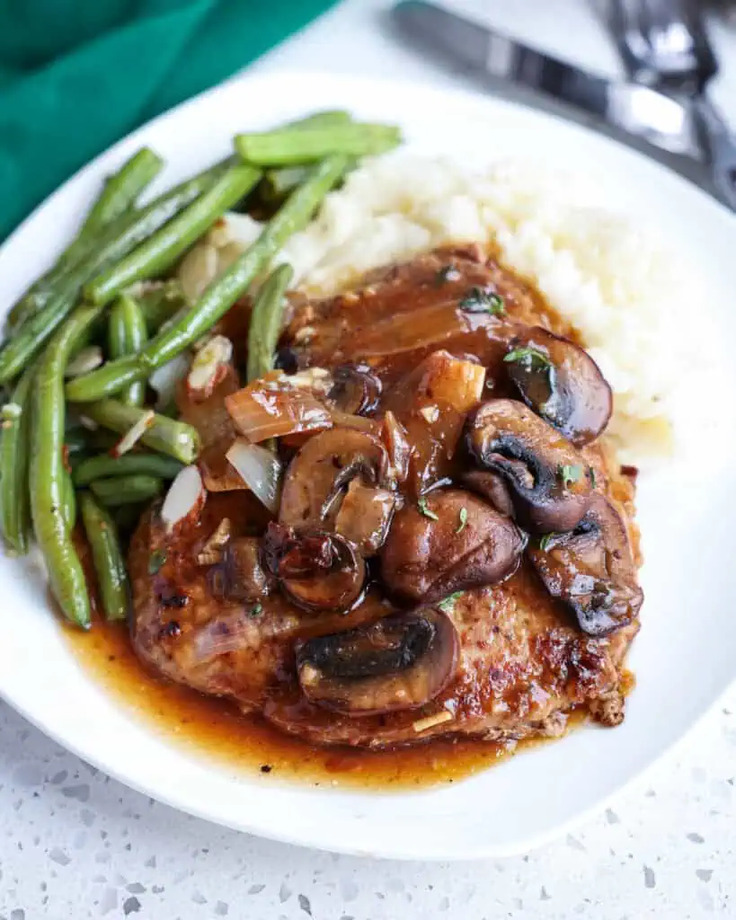 A serving size plate of cube steak, mushrooms, onions, and gravy with mashed potatoes and green beans. 