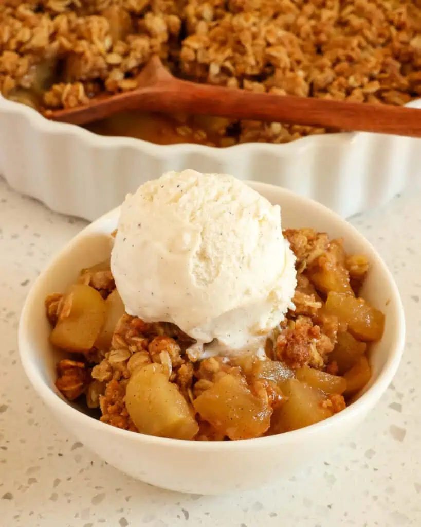 A single serving bowl full of apple crisp topped with a scoop of vanilla ice cream with a baking dish full of apple crisp in the background. 