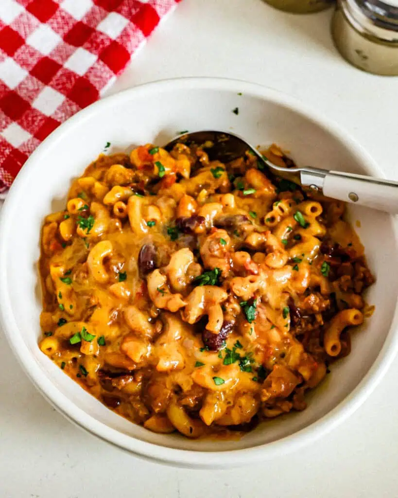 A single serving bowl full cheesy chili mac with a red and white checkered kitchen towel and salt and pepper shaker in the background. 