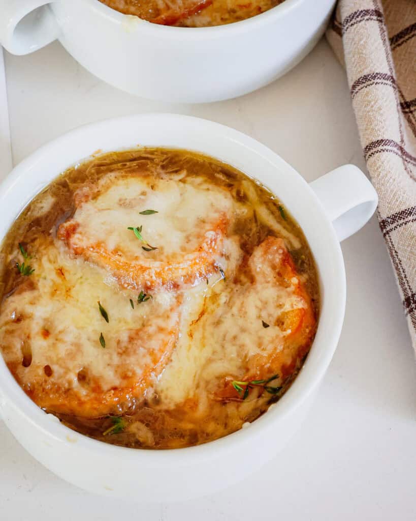 A bowl of French Onion in a soup crock with a brown plain kitchen towel on the side. 