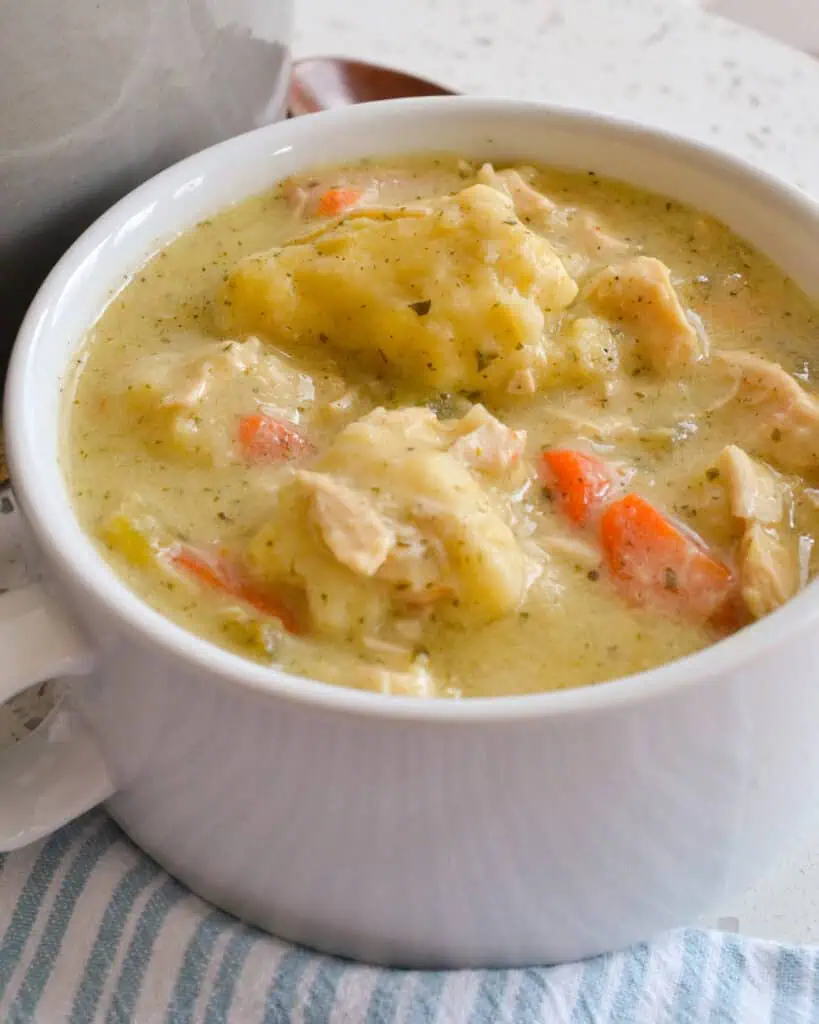 A bowl full of chicken and dumpling soup with a wooden spoon in the background and a blue and white kitchen towel in the foreground. 