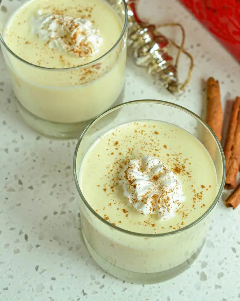 A close up view of two glasses of eggnog with cinnamon sticks and an ornament in the background. 