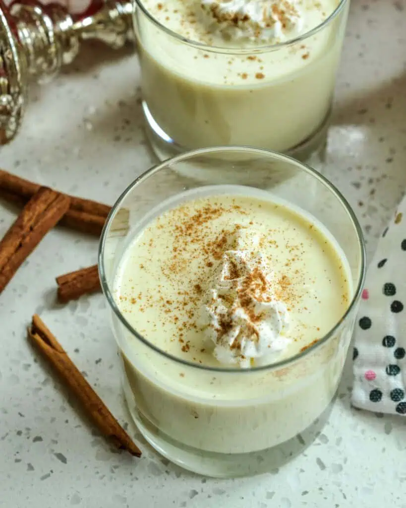 An overhead views of two glasses of eggnog with whipped cream and ground nutmeg.  Cinnamon sticks are on the side and an ornament is in the background. 