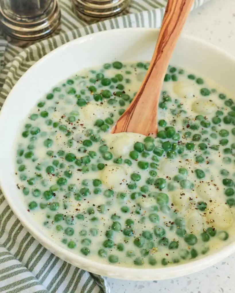 A white serving bowl and woooden spoon full of creamed peas and pearl onions.