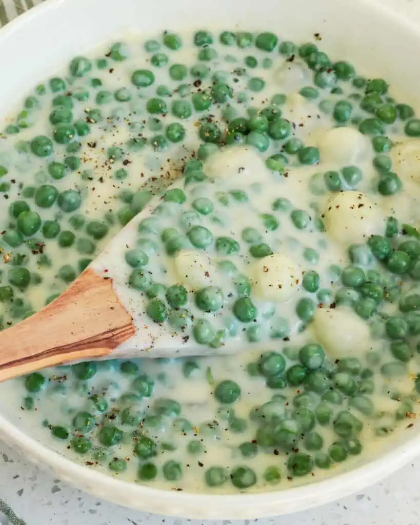 A white round serving bowl and wooden spoon full of creamed peas and pearl onions. 