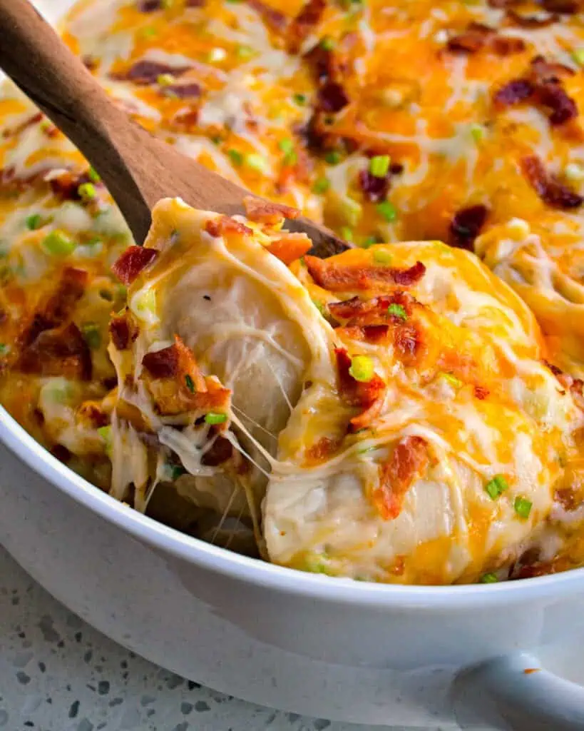 A round casserole dish and a wooden spoon full of pierogi casserole with mashed potato-filled dumplings, alfredo sauce, green onions, bacon, and plenty of shredded mozzarella and cheddar cheese. 