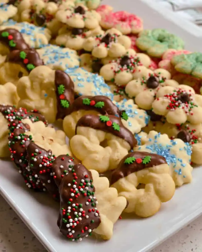 Spritz cookies dipped in chocolate with sprinkles added on a white serving platter. 