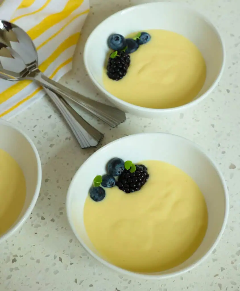 Two bowls of vanilla custard with fresh berries. With spoons and a yellow striped kitchen towel in the background. 