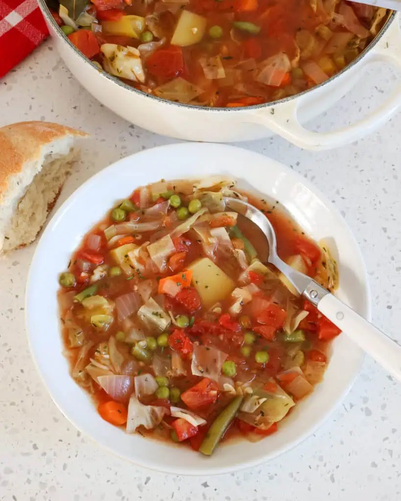 A single serving bowl full of vegetable soup with a piece of crusty French bread on the side and a Dutch oven full of vegetable soup in the bakground. 