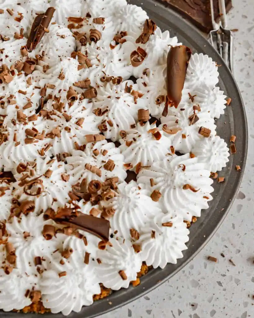 Overhead view of a chocolate cream pie topped with whipped cream and chocolate shavings. There is a peeler and a bar of chocolate in the background. 