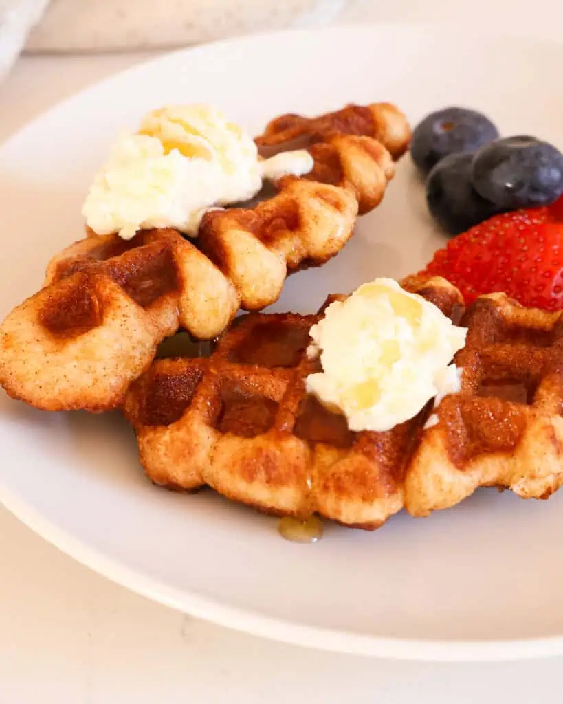 A close up view of two croffles on a plate with butter and syrup. Mixed berries are in the background. 