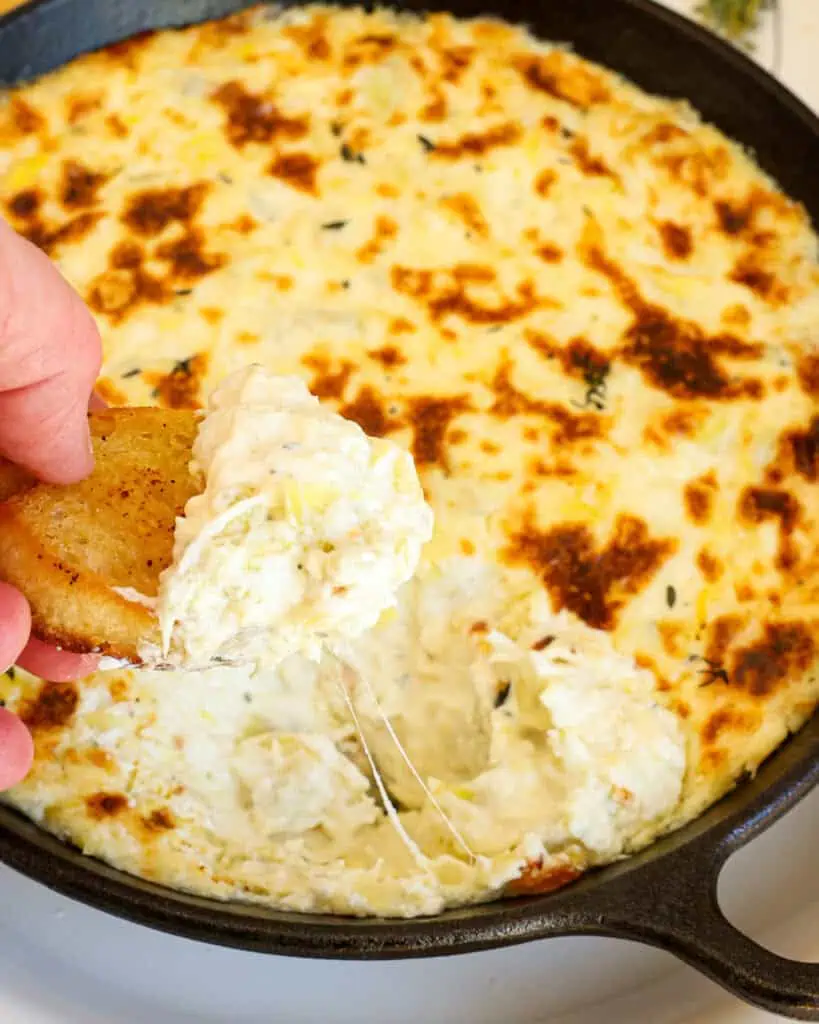 Artichoke dip on a crostini over a small cast iron skillet full of artichoke dip. 