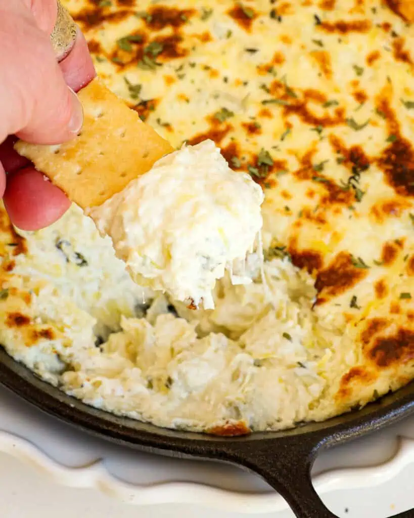 A club cracker full of artihoke dip over a small cast iron skillet full of artichoke dip. 