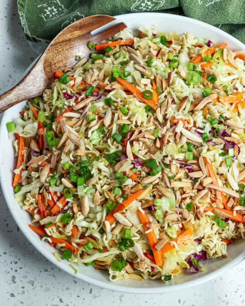 An overhead view of ramen noodle salad in a large round bowl with a wooden serving spoon. 