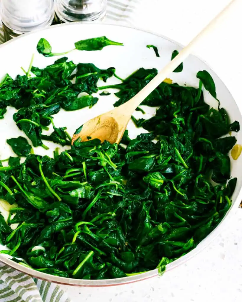 A large skilelt and wooden spoon full of sauteed spinach. 