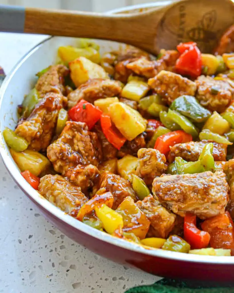 A skillet full of sweet and sour pork with a wooden spoon in the skillet. 