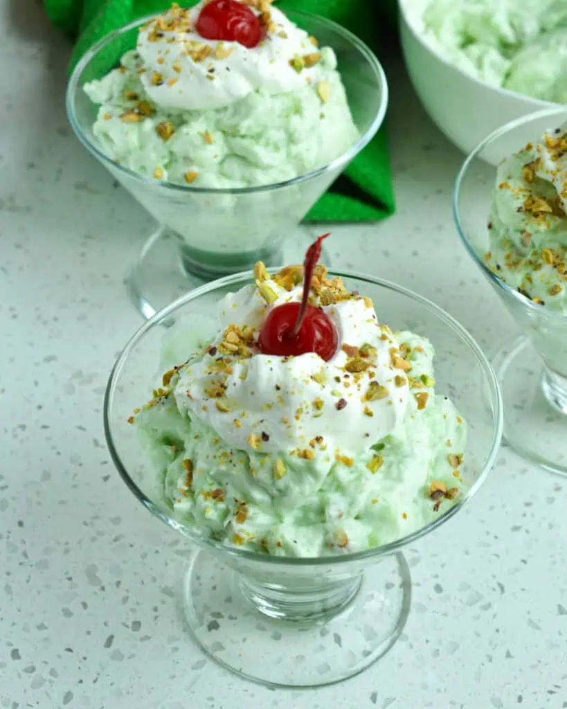 Individual serving dishes full of watergate salad and topped withwhipped cream, chopped pistachios and cherries. 