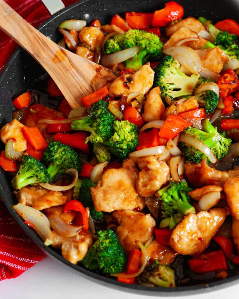 Hunan chicken with brccoli red pepper, and onion in a large skillet with a wooden spatula. 