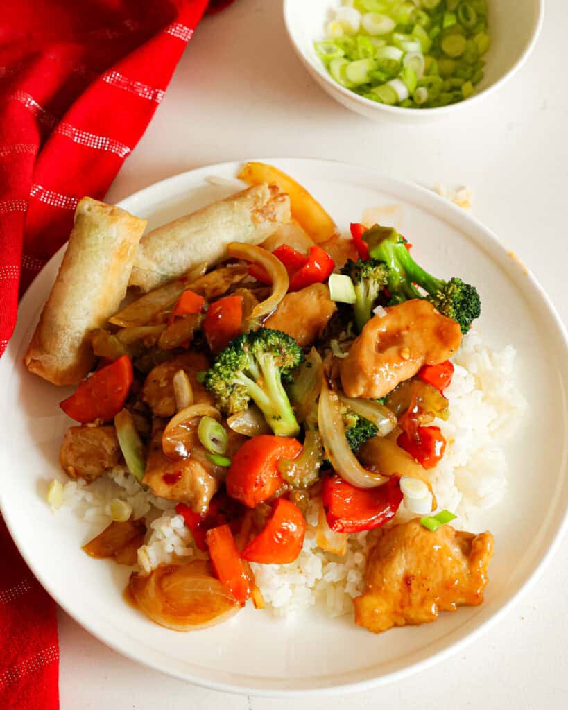 Hunan chicken on a single serving plate with two spring rolls and a bowl of thinly sliced green onions in the bacckground. 