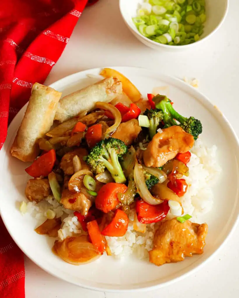 Hunan chicken on a single serving plate with two spring rolls and a bowl of thinly sliced green onions in the bacckground. 