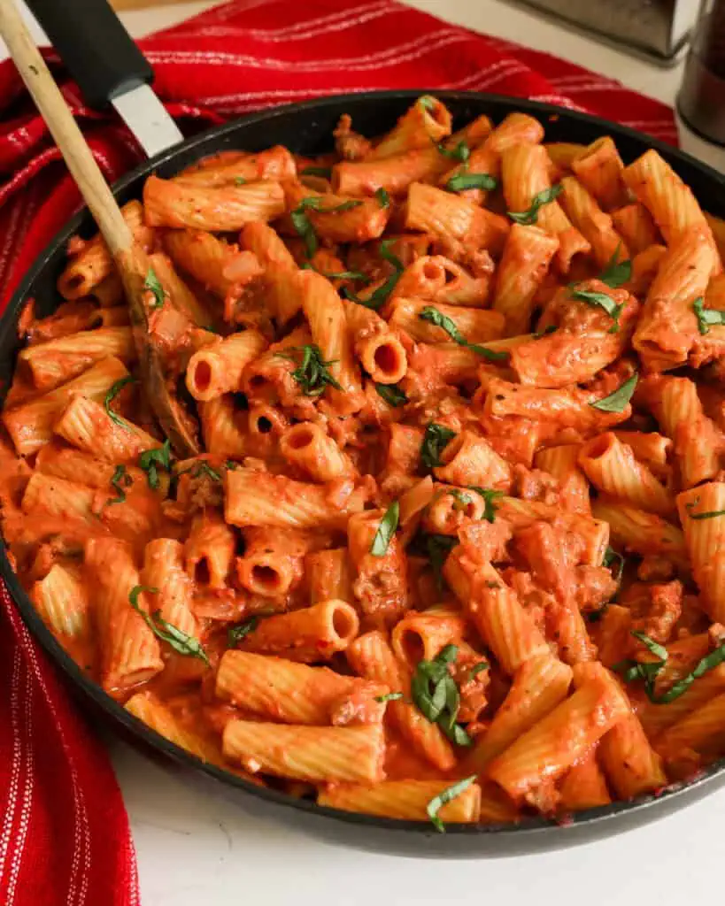 A large skilet full of sausage rigatoni with tomato sauce and fresh basil. 