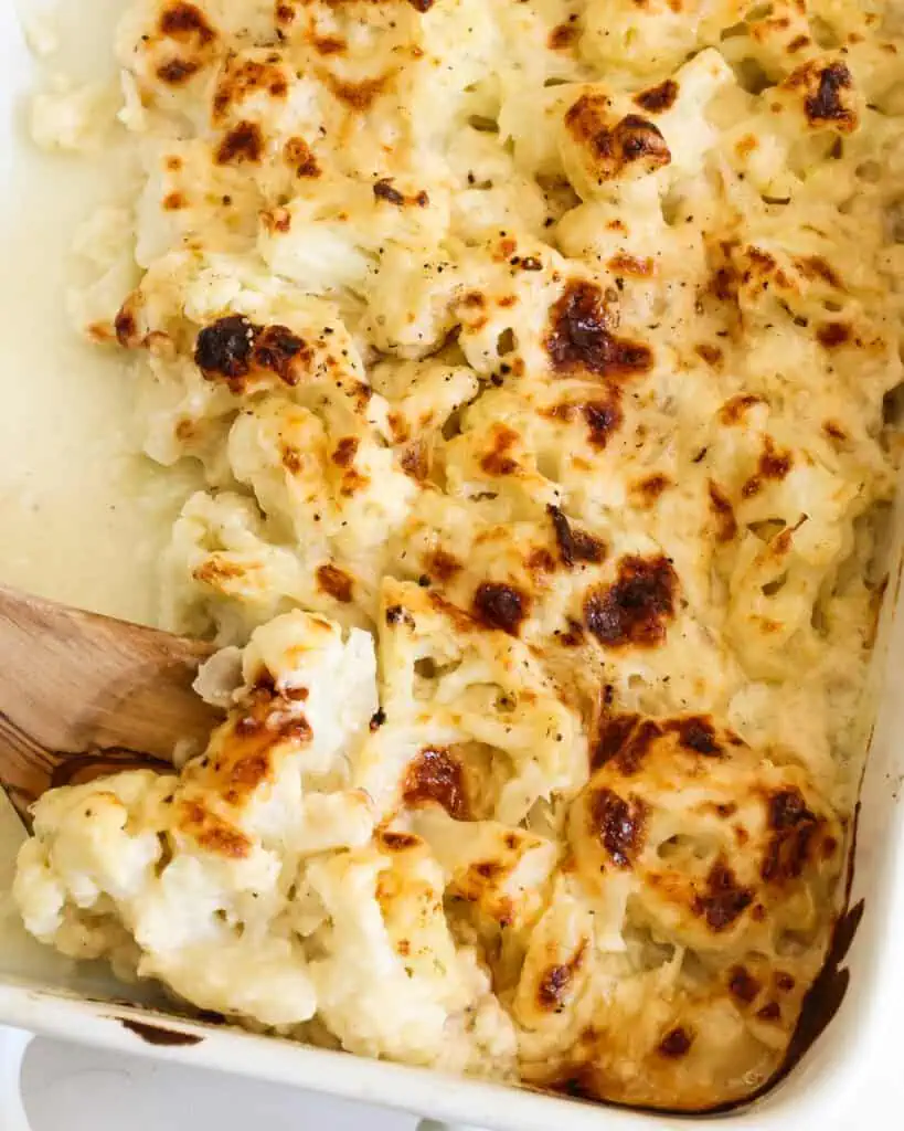 Cheesy cauliflower au gratin in a casserole dish with a large wooden serving spoon. 