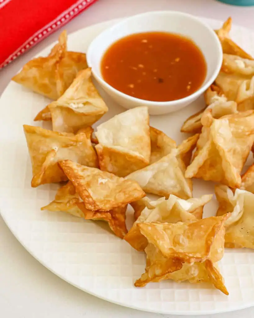 About a dozen cream cheese wontons on a plate with a small bowl of sweet and sour sauce. 